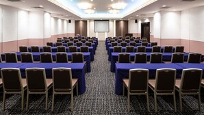 Une salle de conférence avec des rangées de tables et de chaises, un grand écran à l’avant et un plafond orné de lustres.