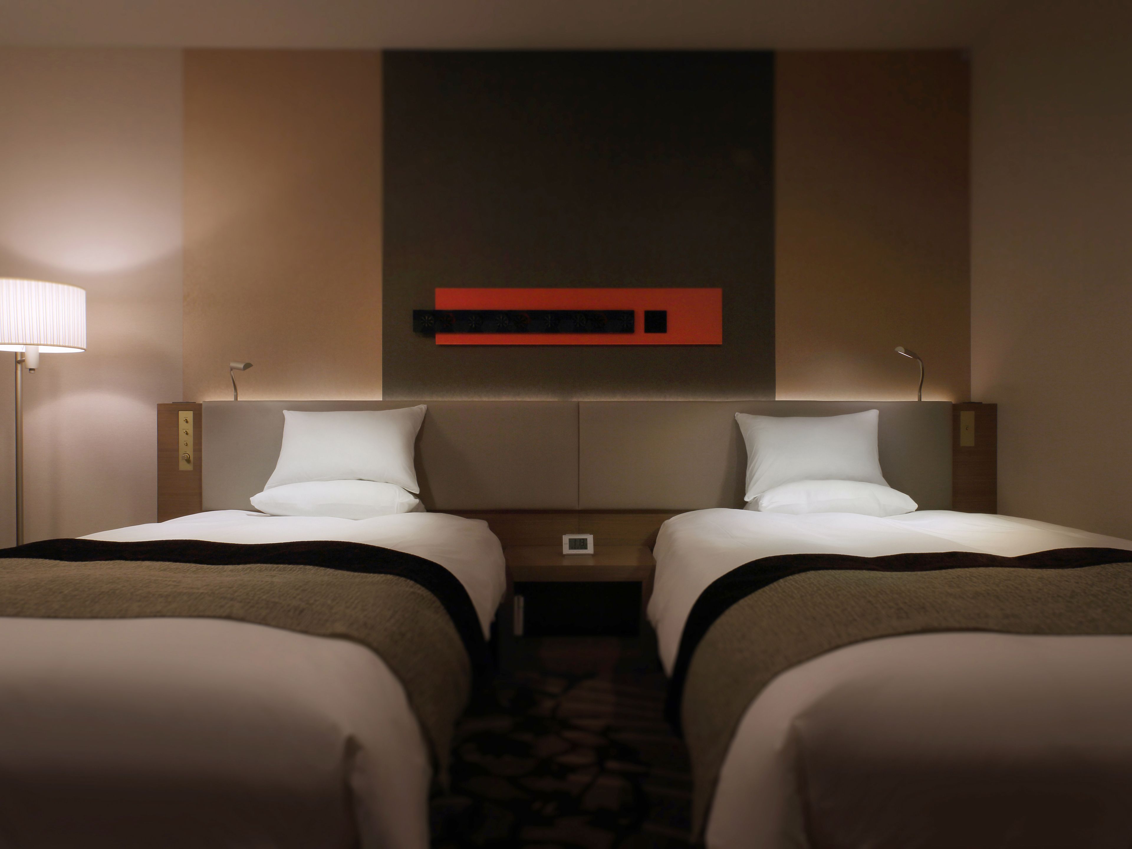 A hotel room with two beds, a nightstand, a lamp, and a wall-mounted control panel.