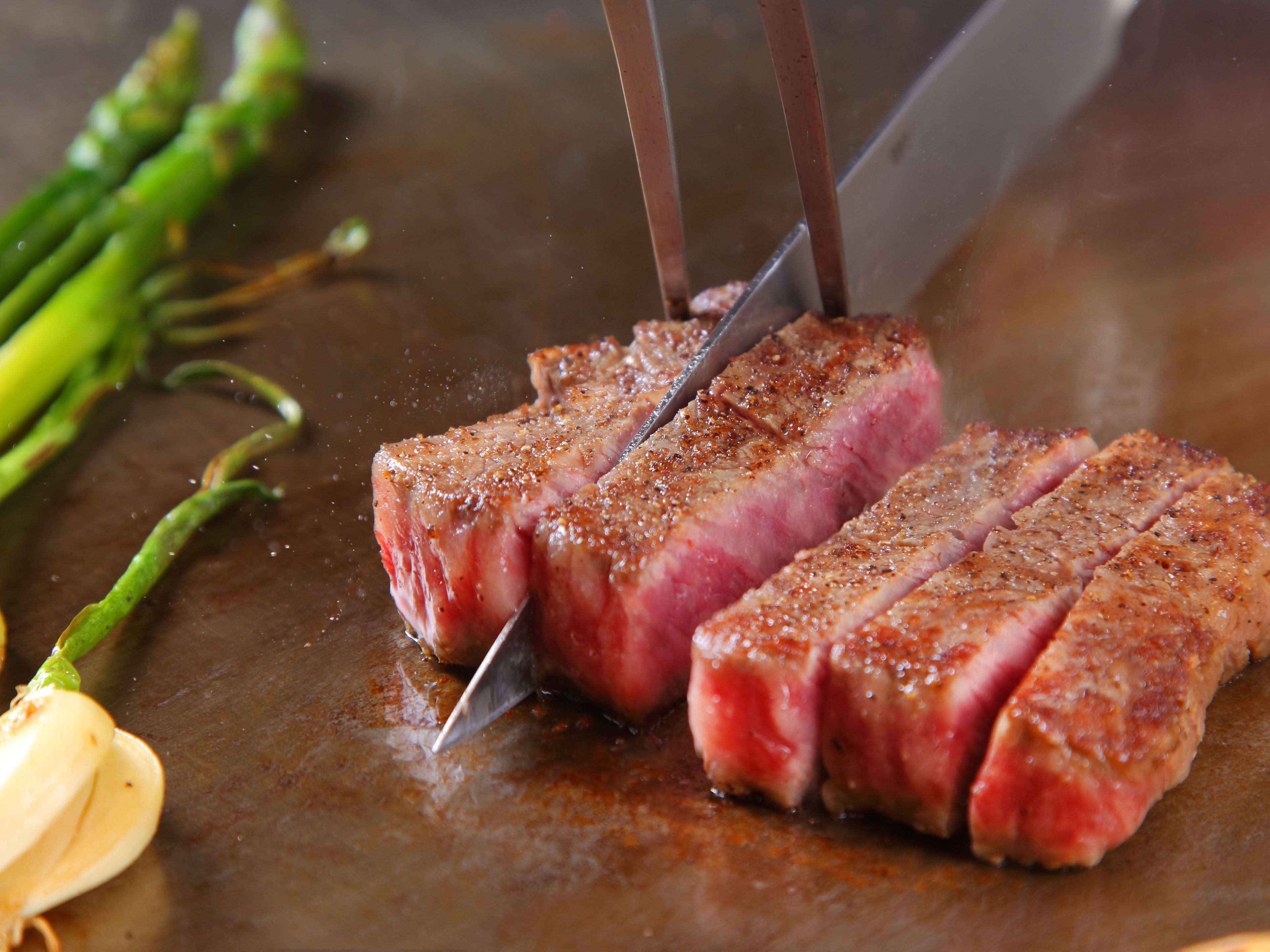 A steak being sliced on a grill with asparagus and garlic nearby.