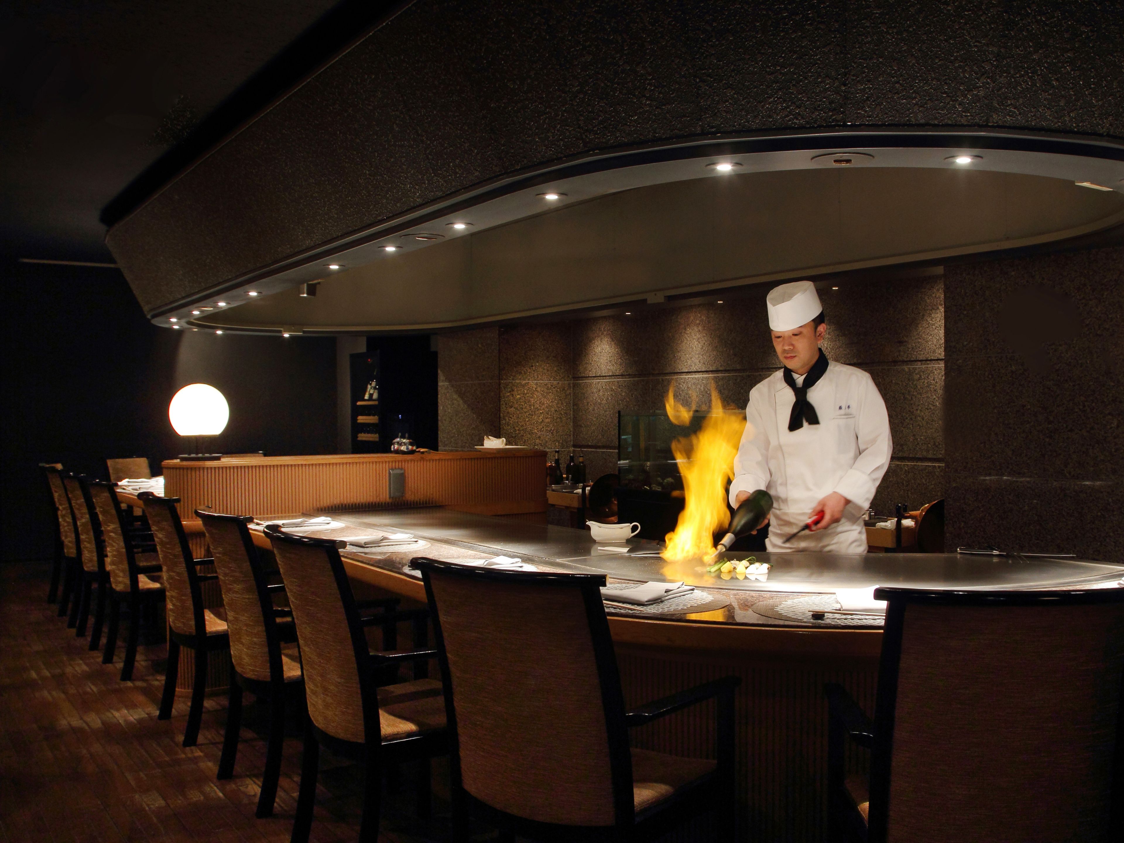 A chef performing a flambé at a restaurant counter with chairs and a bar area in the background.