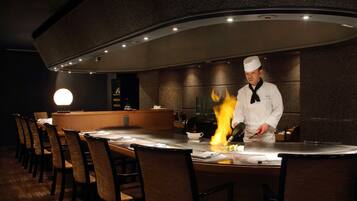 A chef performing a flambé at a restaurant counter with chairs and a bar area in the background.