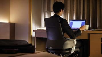 A person sitting at a desk with a laptop, holding a cup, in a room with a bed and a suitcase.