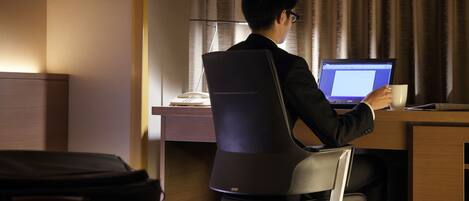 A person sitting at a desk with a laptop, holding a cup, in a room with a bed and a suitcase.