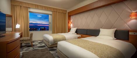 A hotel room with two beds, a desk, a chair, a TV, and a view of the cityscape at dusk.