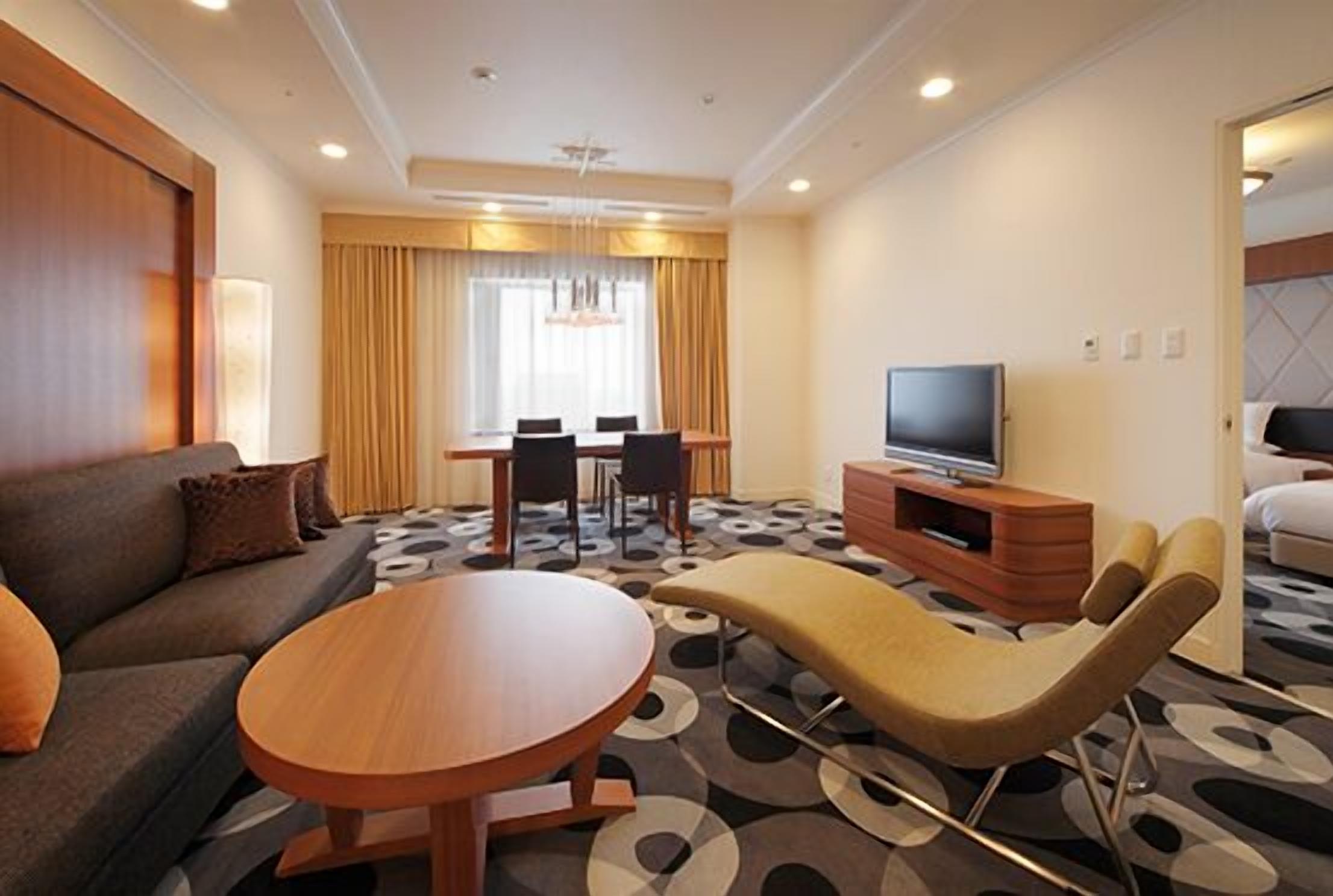 Una habitación de hotel moderna con sofá, una mesa redonda, televisor y un área de comedor con sillas y una mesa.