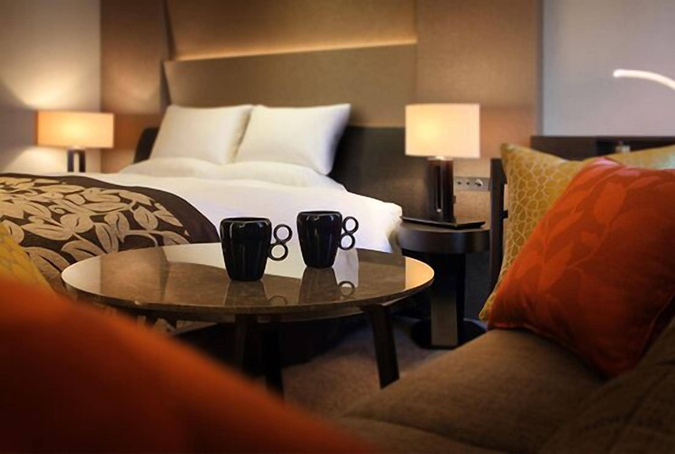 A hotel room with a bed, two bedside lamps, a round table with two black mugs, and a sofa with a patterned throw.