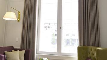 Una habitación de hotel con una cama, un sillón verde, un sofá morado, una mesita con un jarrón de flores y un ventanal con cortinas.
