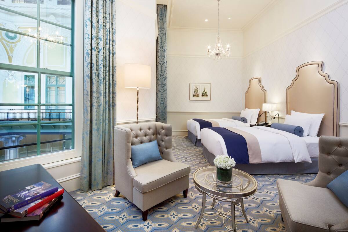Habitación de hotel con un ventanal grande, dos camas, un área de descanso con una silla, una mesita con un florero y una araña.
