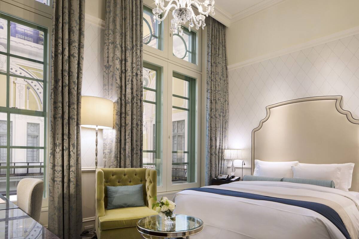 Habitación de hotel con una cama grande, una zona de estar con una silla, una mesa redonda de vidrio y una araña.