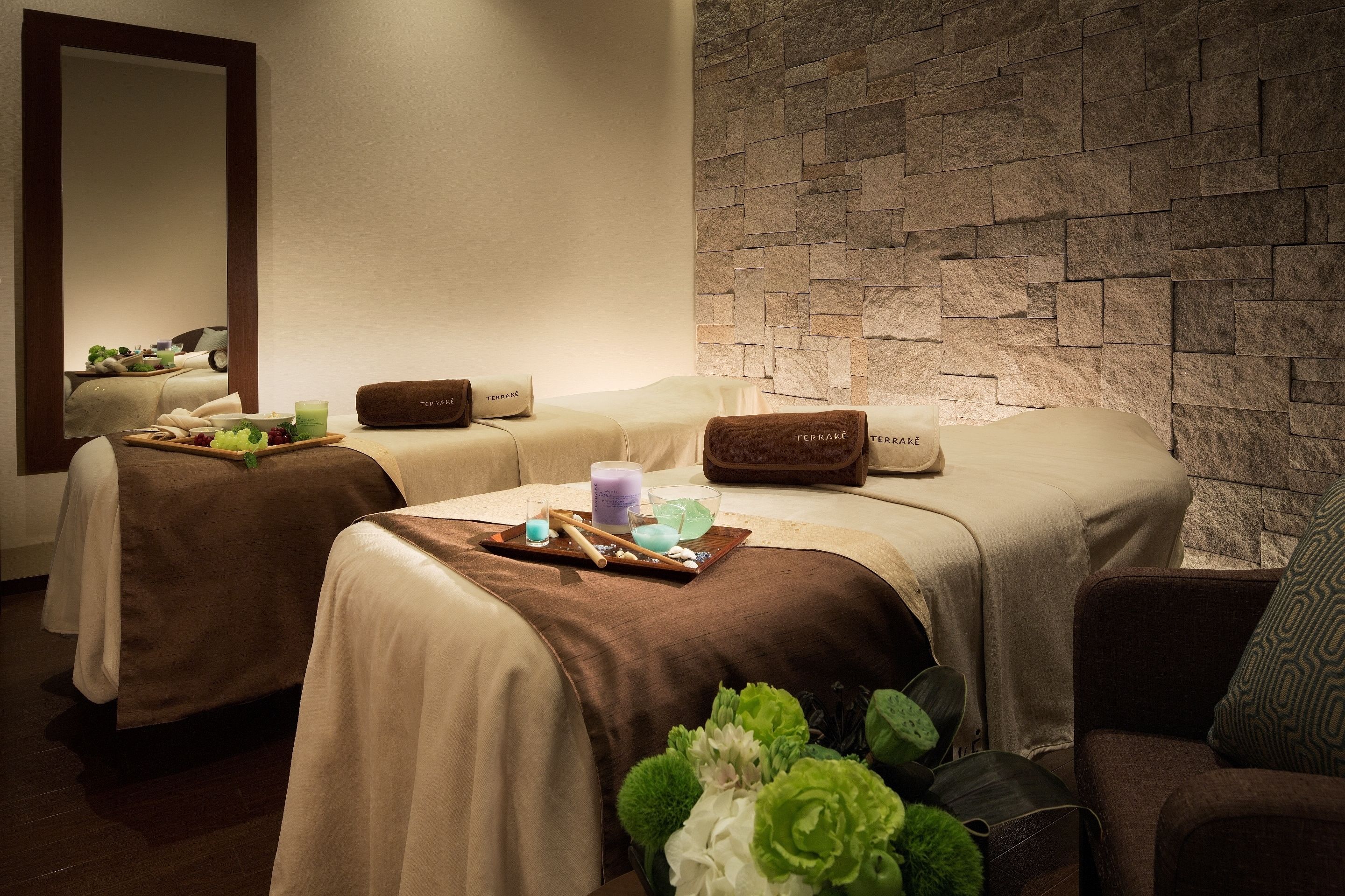 A spa room with two massage beds, a stone wall, and a mirror reflecting a vehicle.