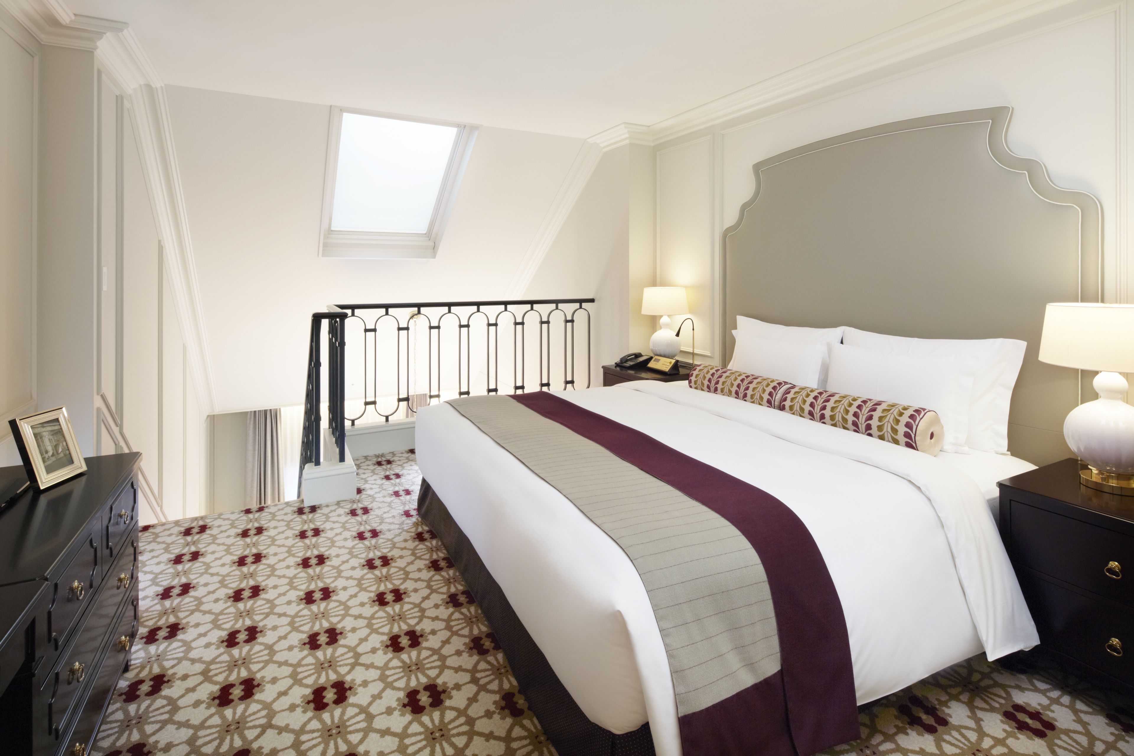 A hotel room with a large bed, a nightstand with lamps, a dresser, and a patterned carpet.