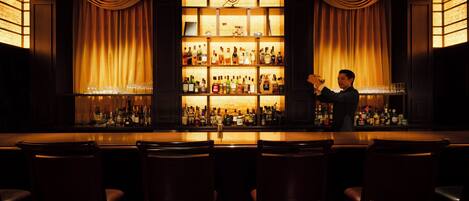 Zona de bar con un bartender, estantes con botellas de licores y taburetes.