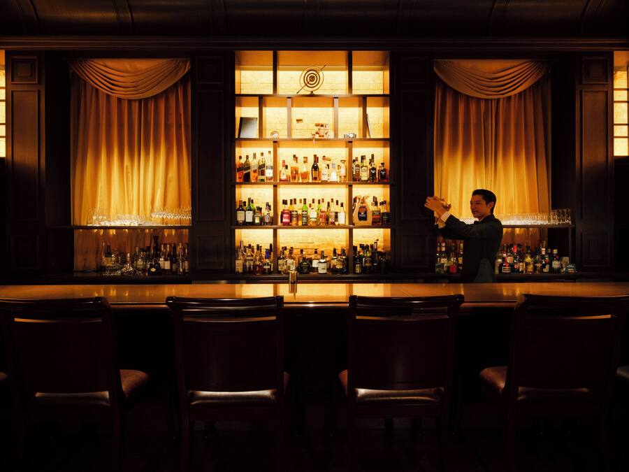 A bar area with a bartender, shelves of liquor bottles, and bar stools.