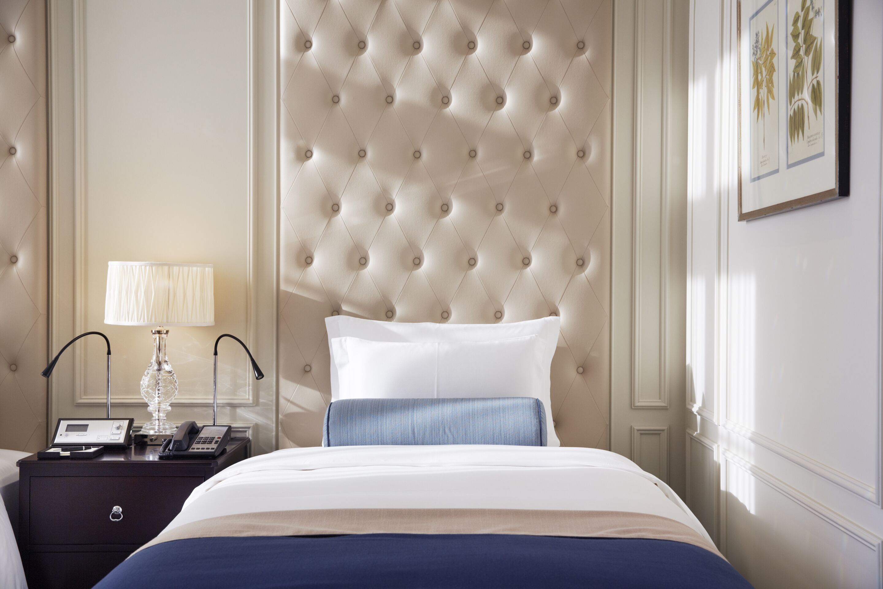 Una habitación de hotel con cabecera capitonada, una mesita de noche con lámpara, teléfono e impresión botánica enmarcada en la pared.