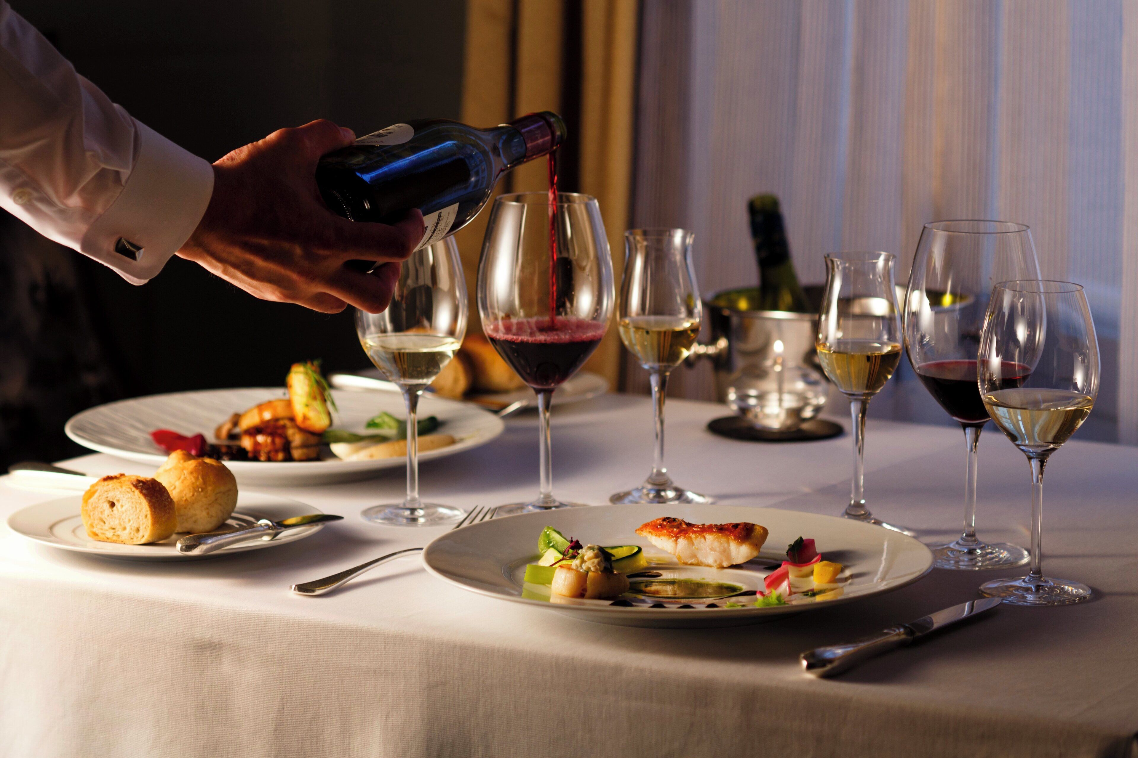 A dining table with plates of food, wine glasses, and a bottle being poured.