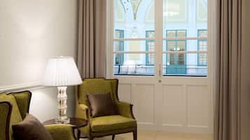 A room with a large window, a chandelier, two green armchairs, a side table, and a patterned rug.