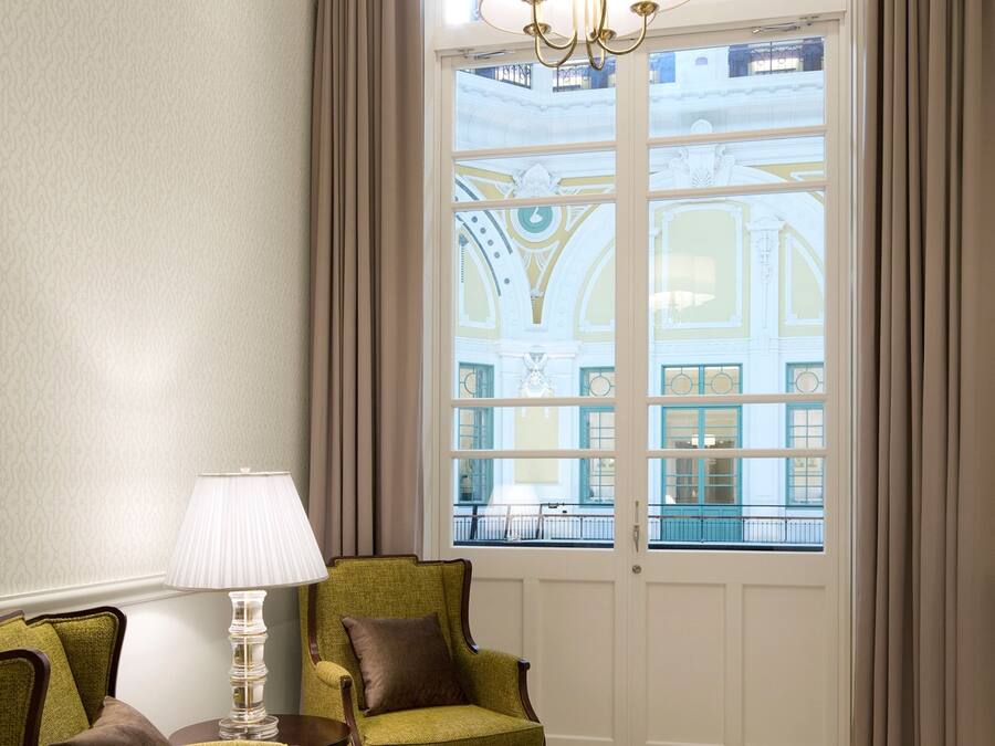 A room with a large window, a chandelier, two green armchairs, a side table, and a patterned rug.