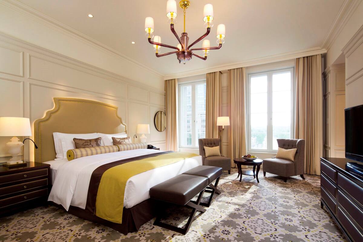 Habitación de hotel con una cama grande, un banco, dos sillones, una mesita, un televisor y un candelabro.