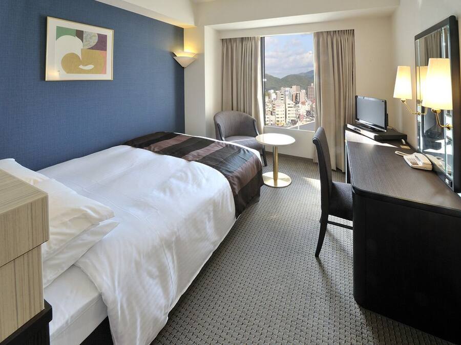 A hotel room with a bed, a desk with a chair, a television, and a window with a city view.
