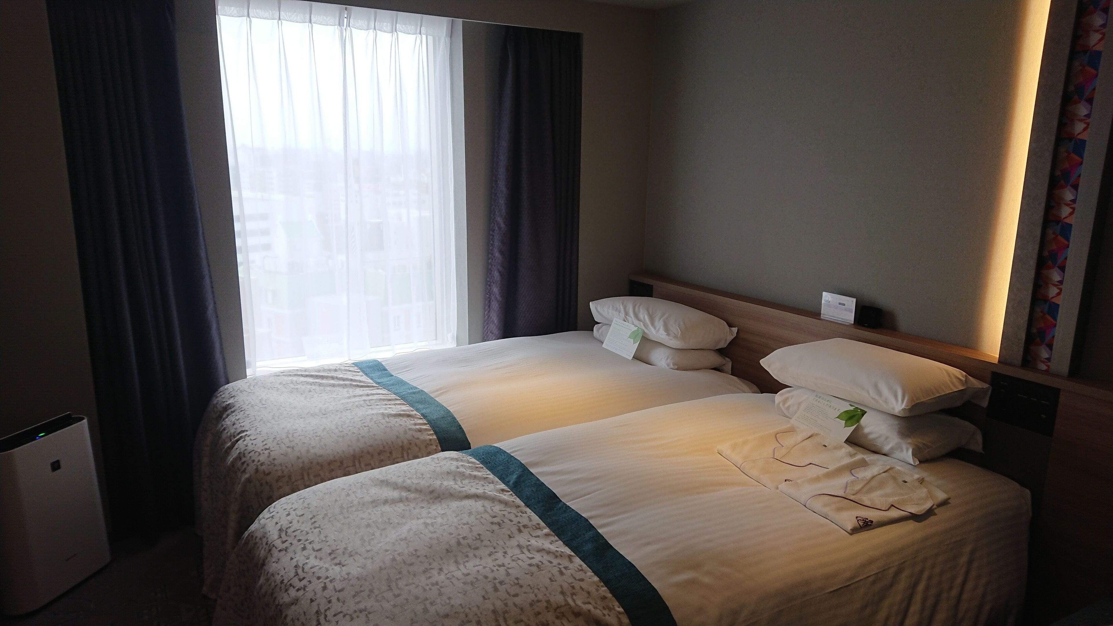 A hotel room with two beds, a window with curtains, a bedside table, and a wall-mounted device.