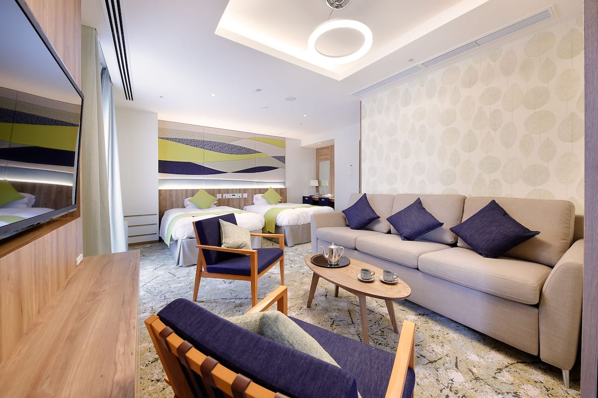 A modern hotel room with a sofa, two armchairs, a coffee table, and a bed with a geometric-patterned headboard.