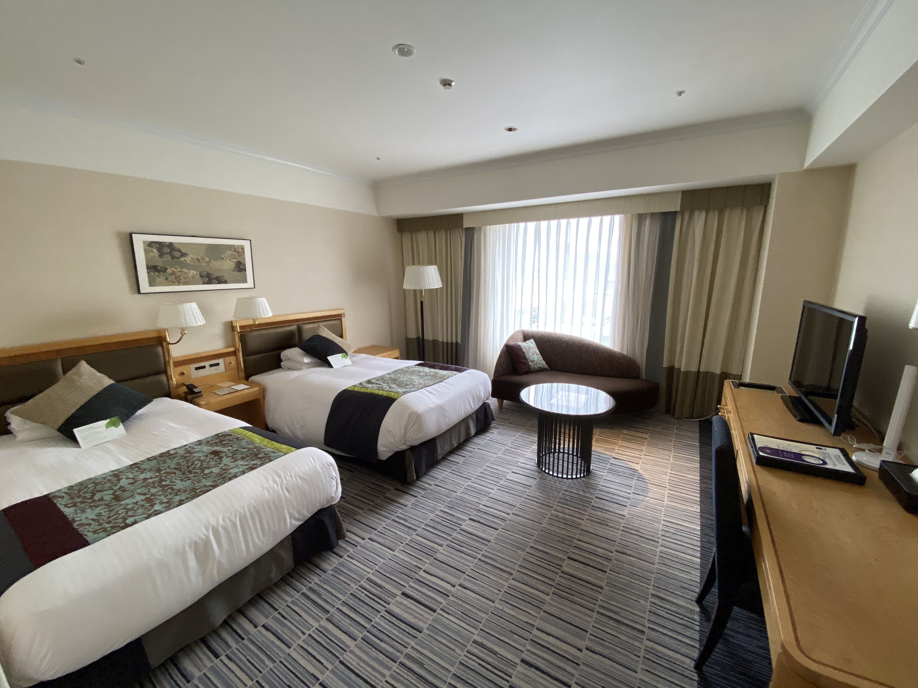 A hotel room with two beds, a sofa, a small table, a TV, and a window with curtains.