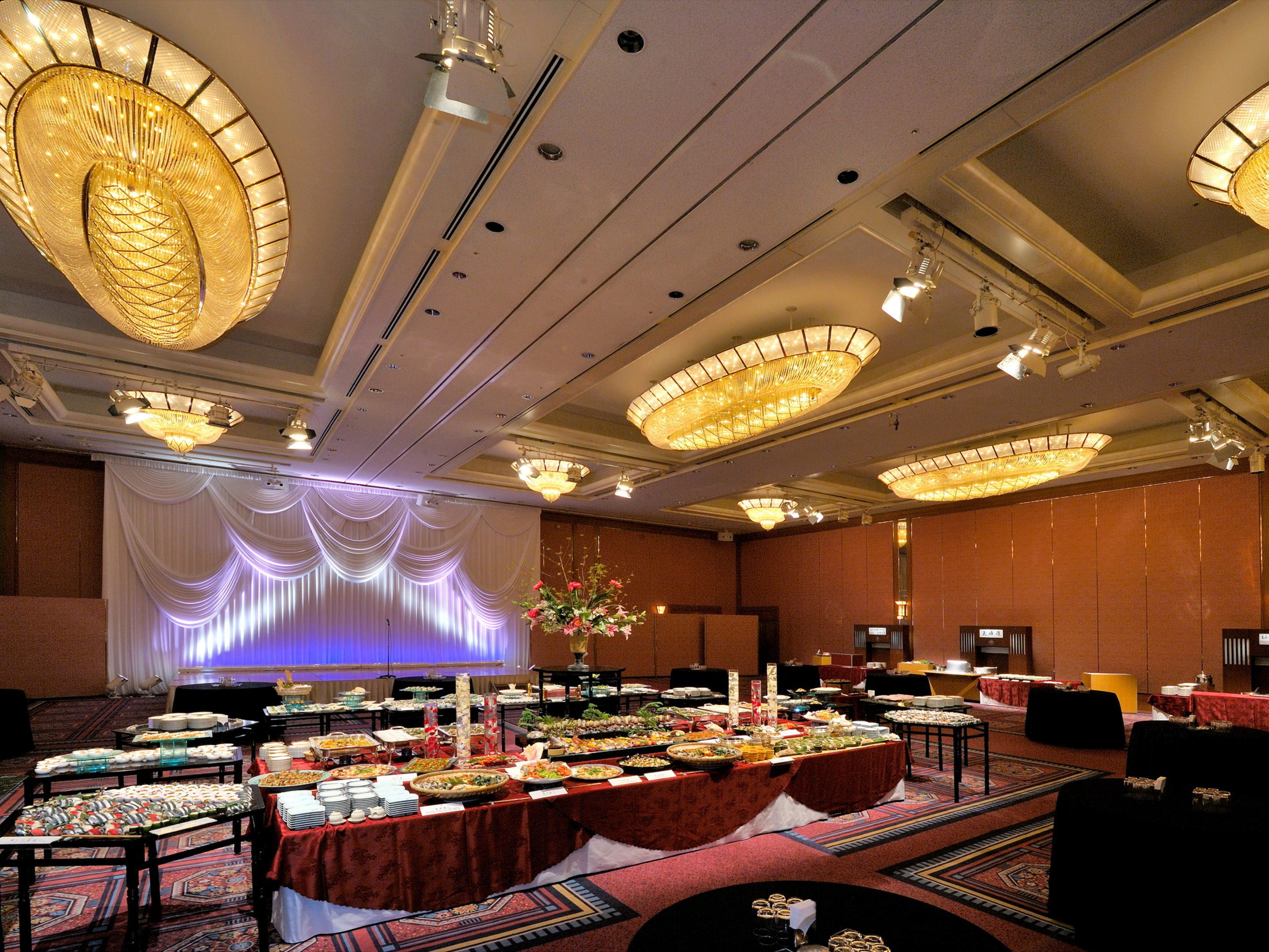 A banquet hall with multiple tables covered with food, large chandeliers, and a stage with draped curtains.