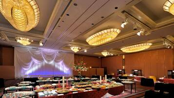A banquet hall with multiple tables covered with food, large chandeliers, and a stage with draped curtains.