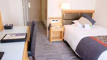 A hotel room with a bed, bedside table, lamp, and a desk with a chair.
