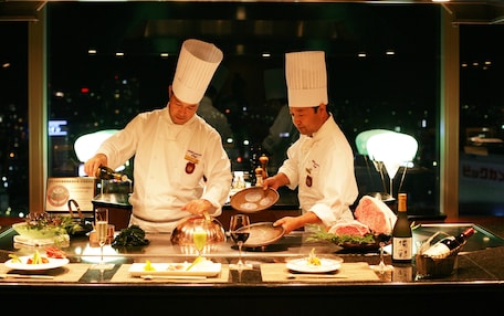 Two chefs in white uniforms preparing food at a counter with various dishes and drinks.