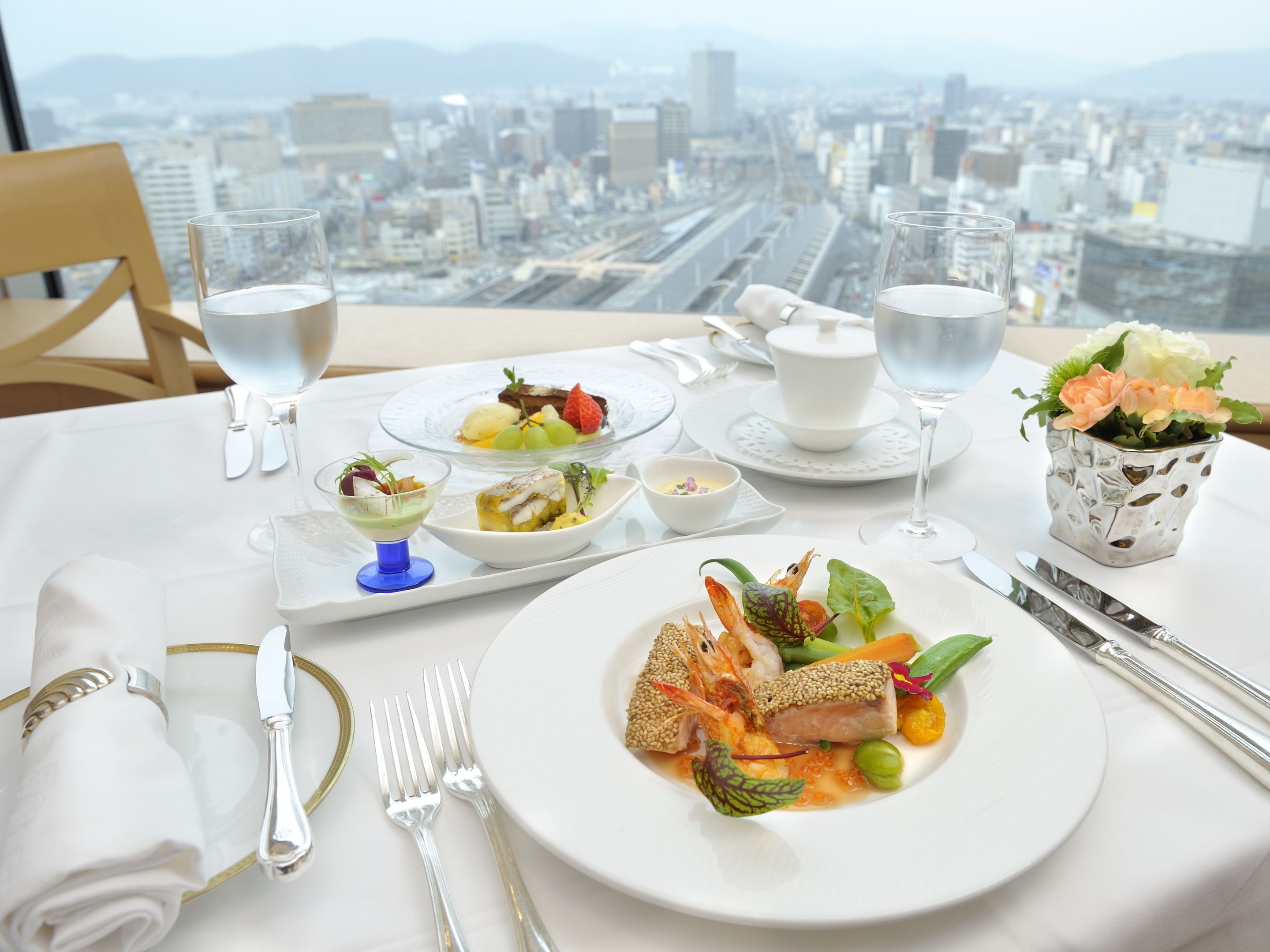 Mesa de comedor con vista a la ciudad, mantel blanco y diversos platos.