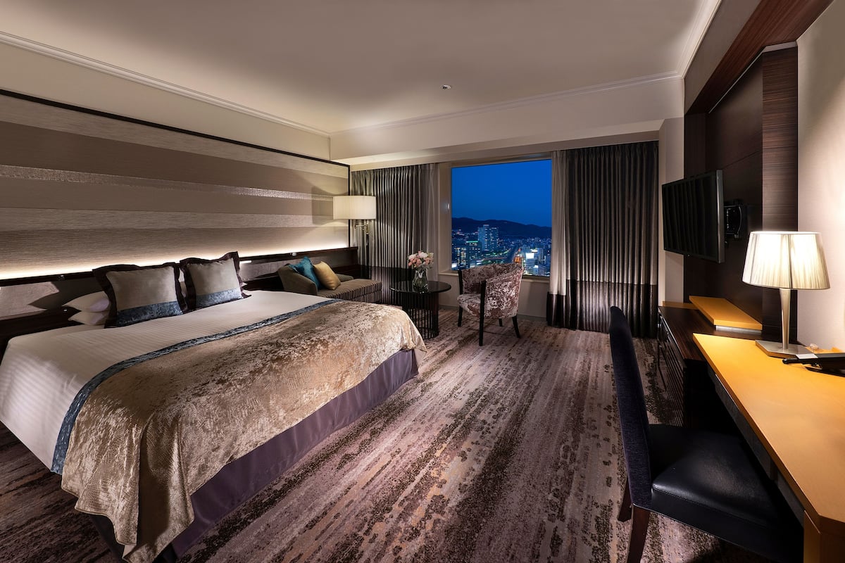 A modern hotel room with a large bed, a desk, a chair, and a view of the city at night.