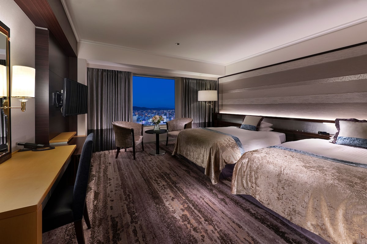 A hotel room with a large bed, a desk, and a chair. There is a TV mounted on the wall and a window with a view of the city.