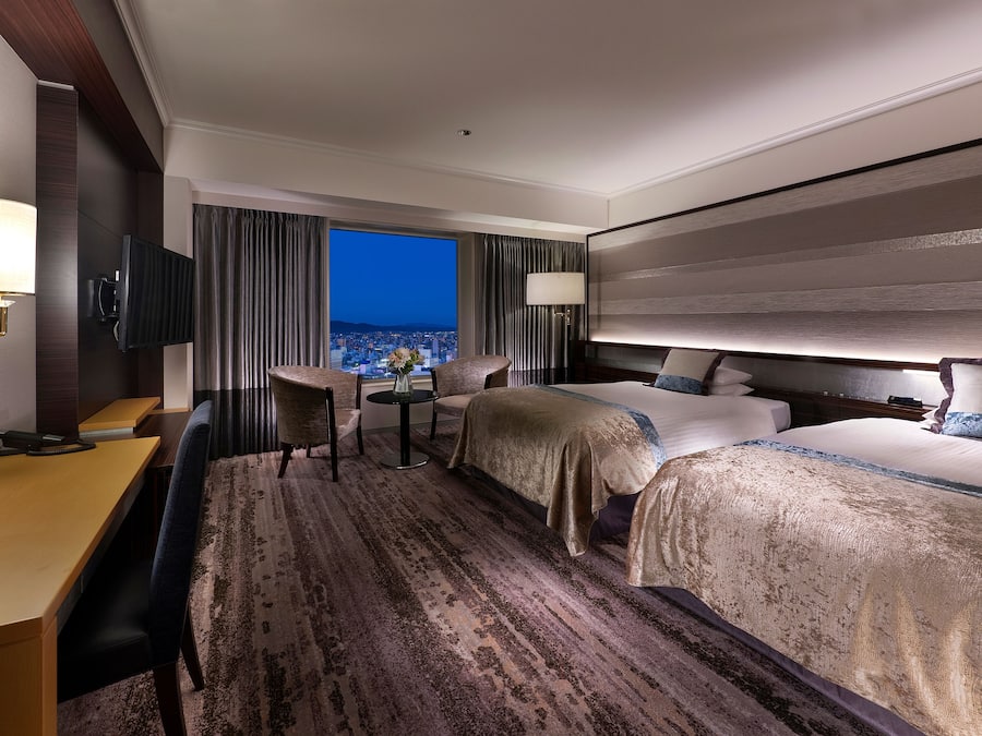 Habitación de hotel con una cama grande, un escritorio y una silla. Hay un televisor montado en la pared y una ventana con vistas a la ciudad.