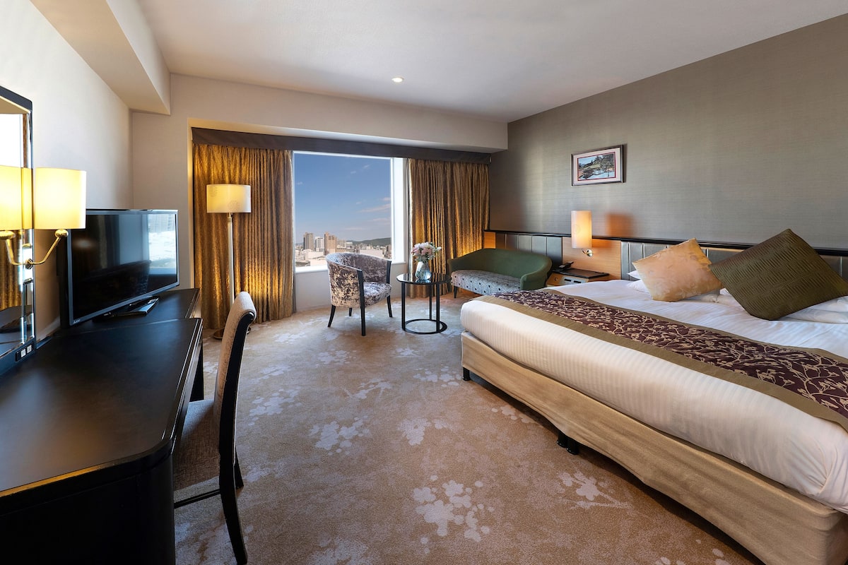 A hotel room with a large bed, a desk with a TV, a chair, and a view of the city through the window.