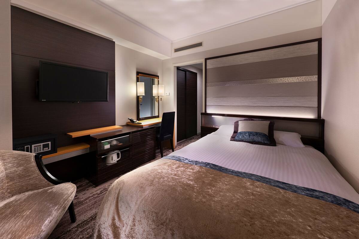 A hotel room with a bed, a desk with a computer, a chair, a television mounted on the wall, and a large mirror.