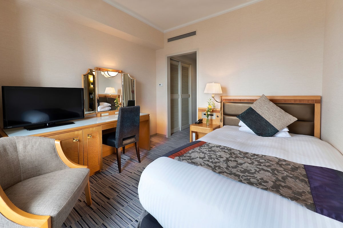 A hotel room with a bed, a desk with a television, a chair, a nightstand with a lamp, and a mirror.