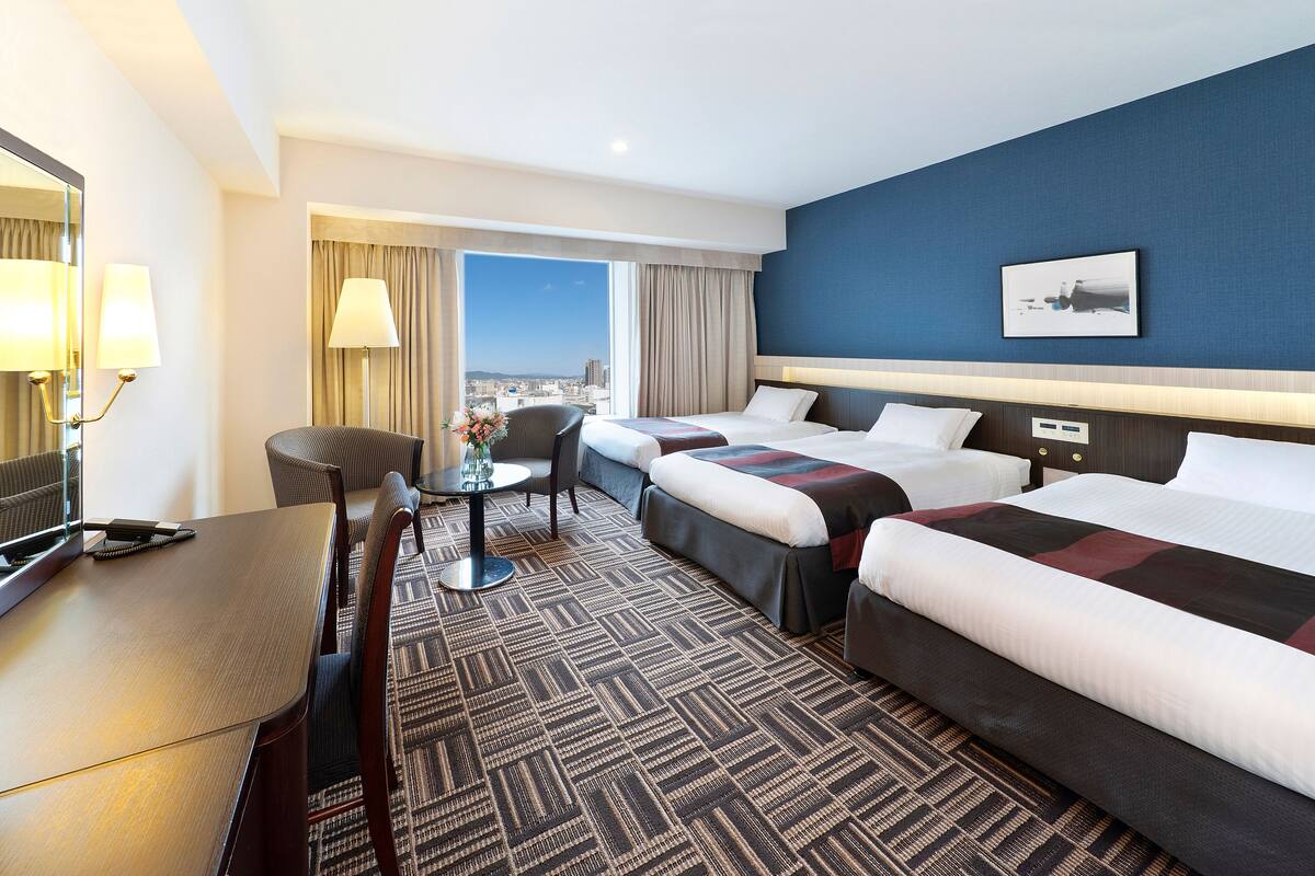 A hotel room with two beds, a desk, a chair, a table, and a view of the city.