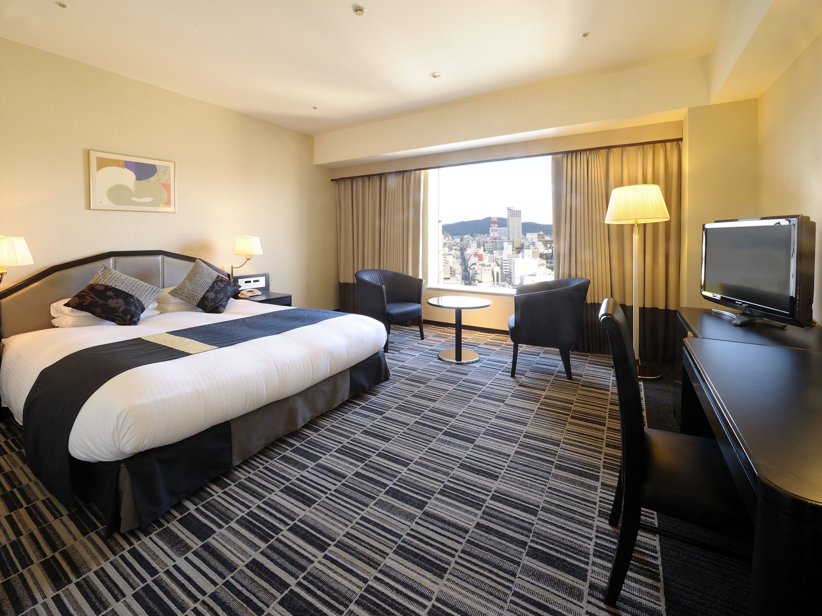 A hotel room with a large bed, a desk with a television, two chairs, a small table, and a view of a cityscape through the window.