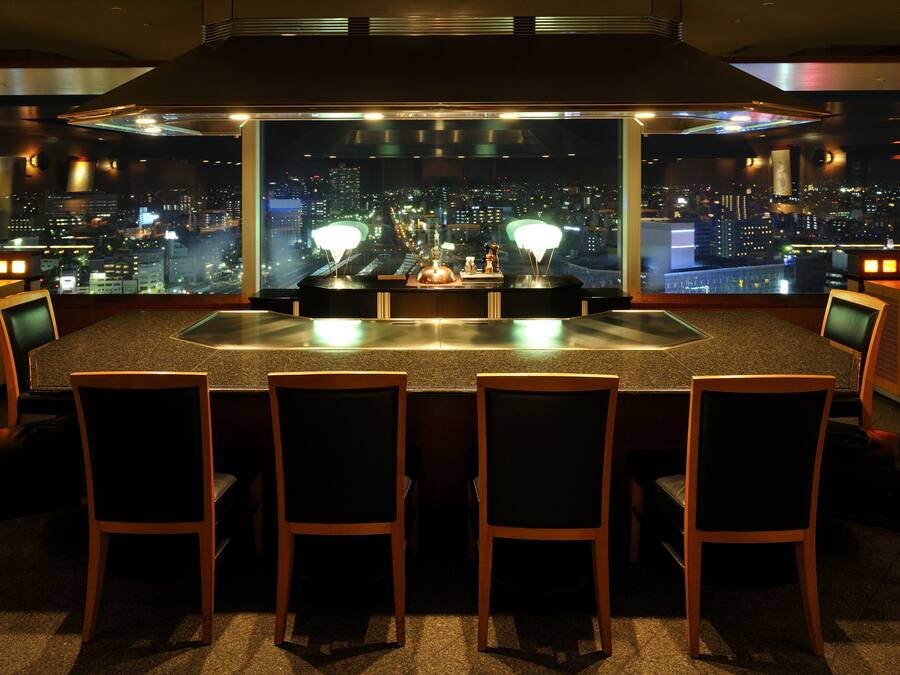 Zona de bar con vista a la ciudad, varias sillas y una mesa central con una lámpara.