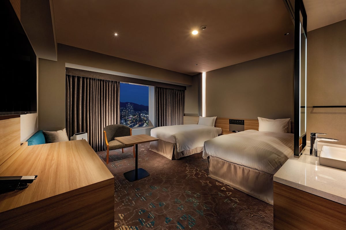 A hotel room with two beds, a desk, a chair, a large window with curtains, and a city view.