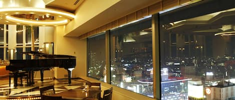 A high-rise hotel room with a city view, a grand piano, and a dining area with a table and chairs.