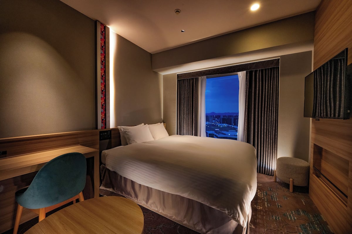 A hotel room with a bed, a desk, a chair, a small stool, and a large window with curtains.