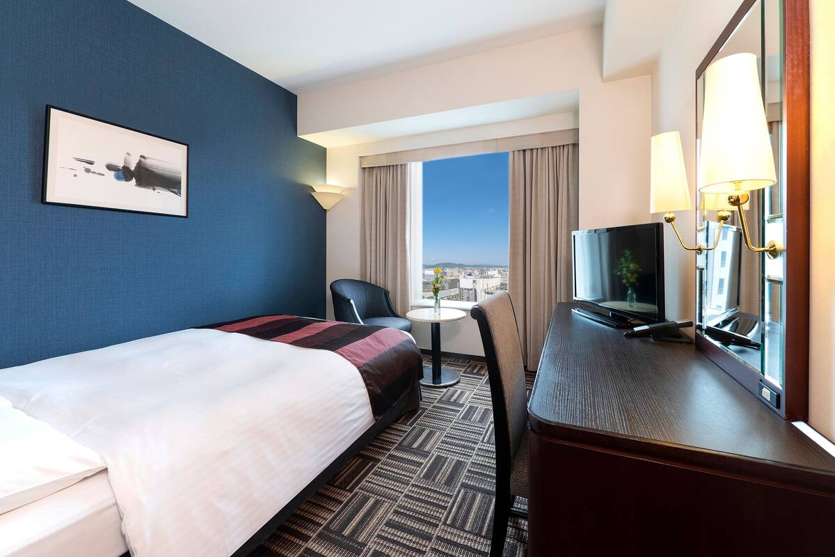 A hotel room with a bed, a desk with a TV, a chair, a small table, and a window with a city view.