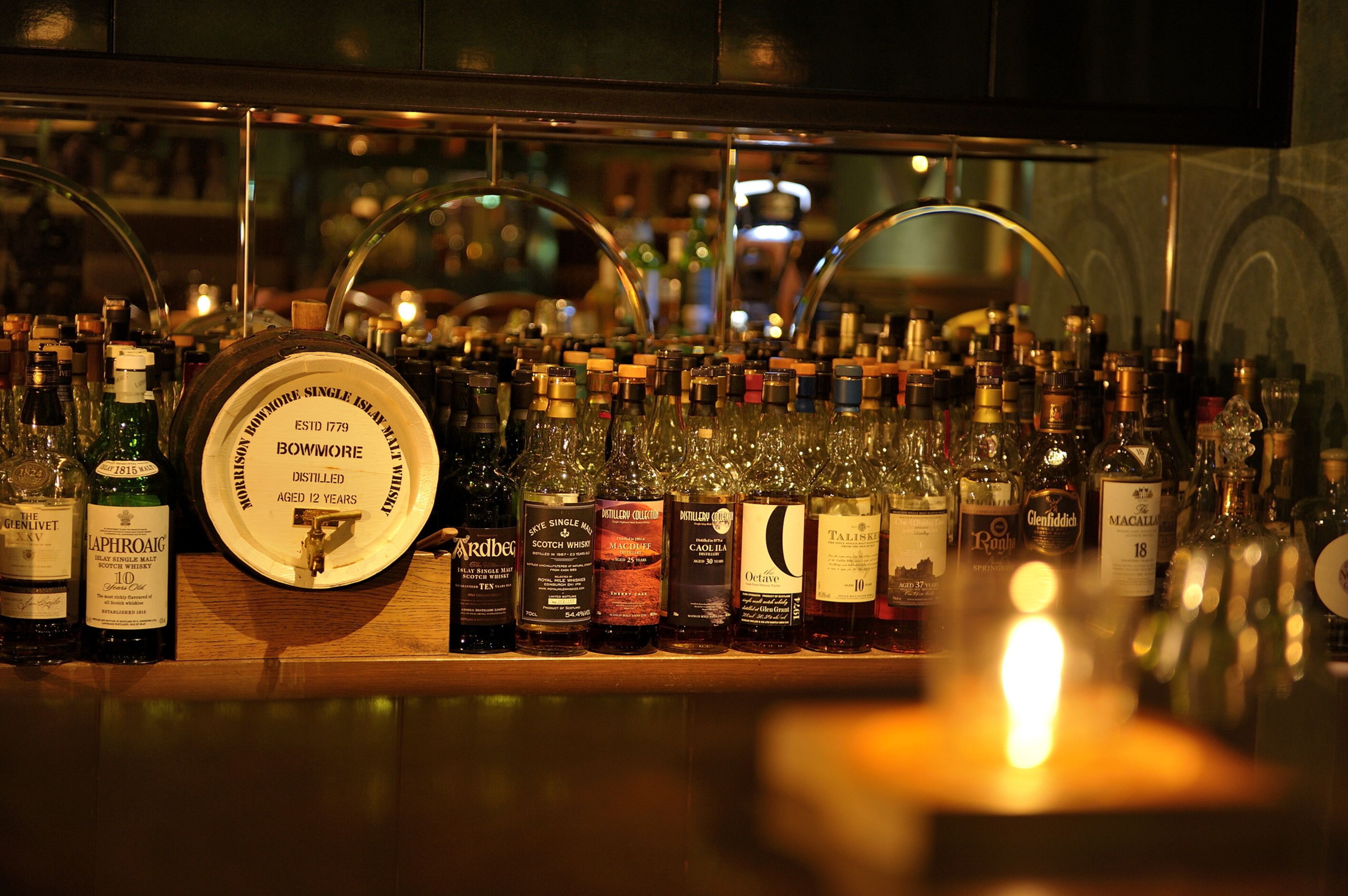 Un bar con una variedad de botellas y vasos, incluyendo una botella redonda grande con la etiqueta "BOWMORE SINGLE ISLAND HILL ESTD 1779".