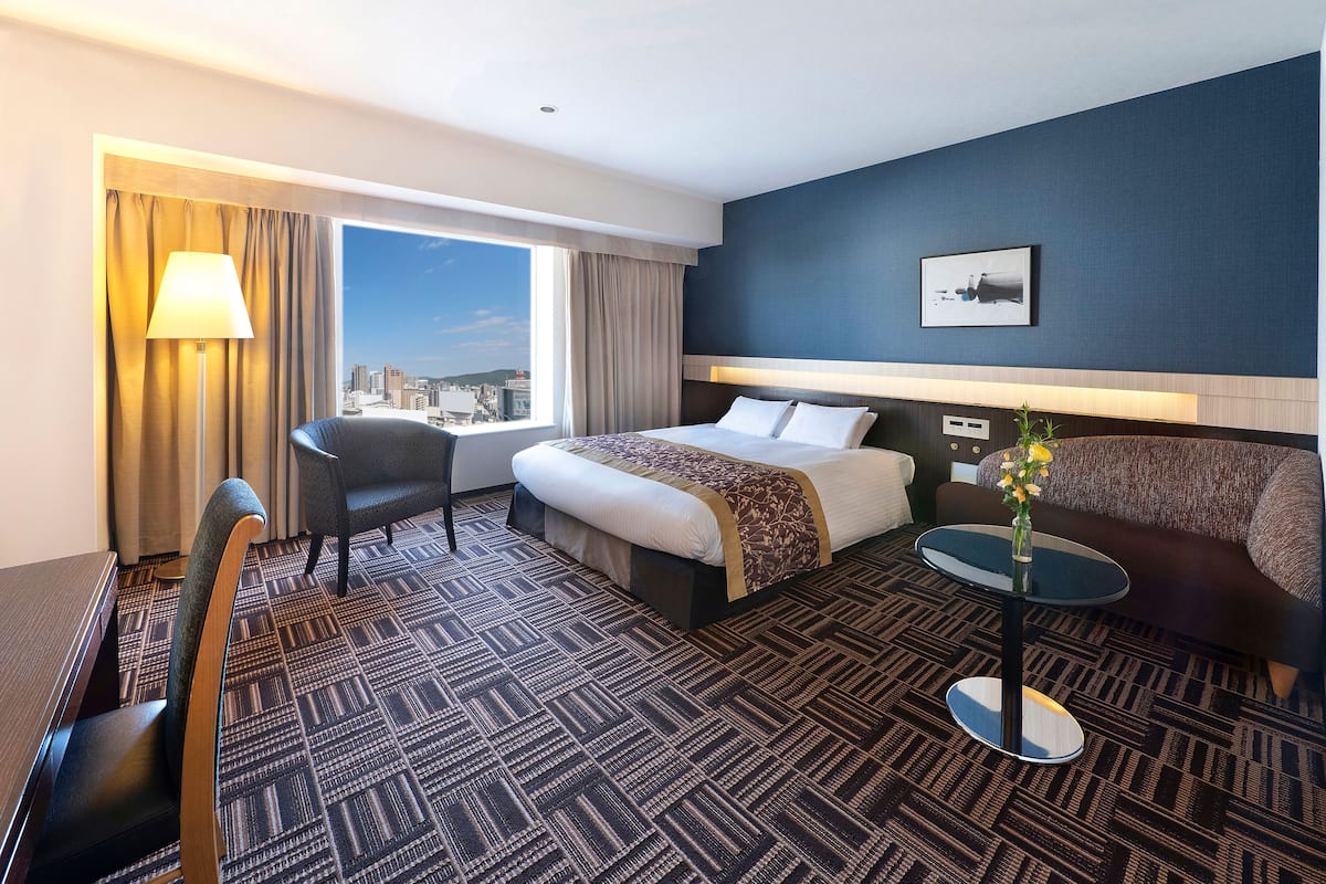 A hotel room with a bed, a sofa, a desk, a chair, a lamp, and a view of the city.