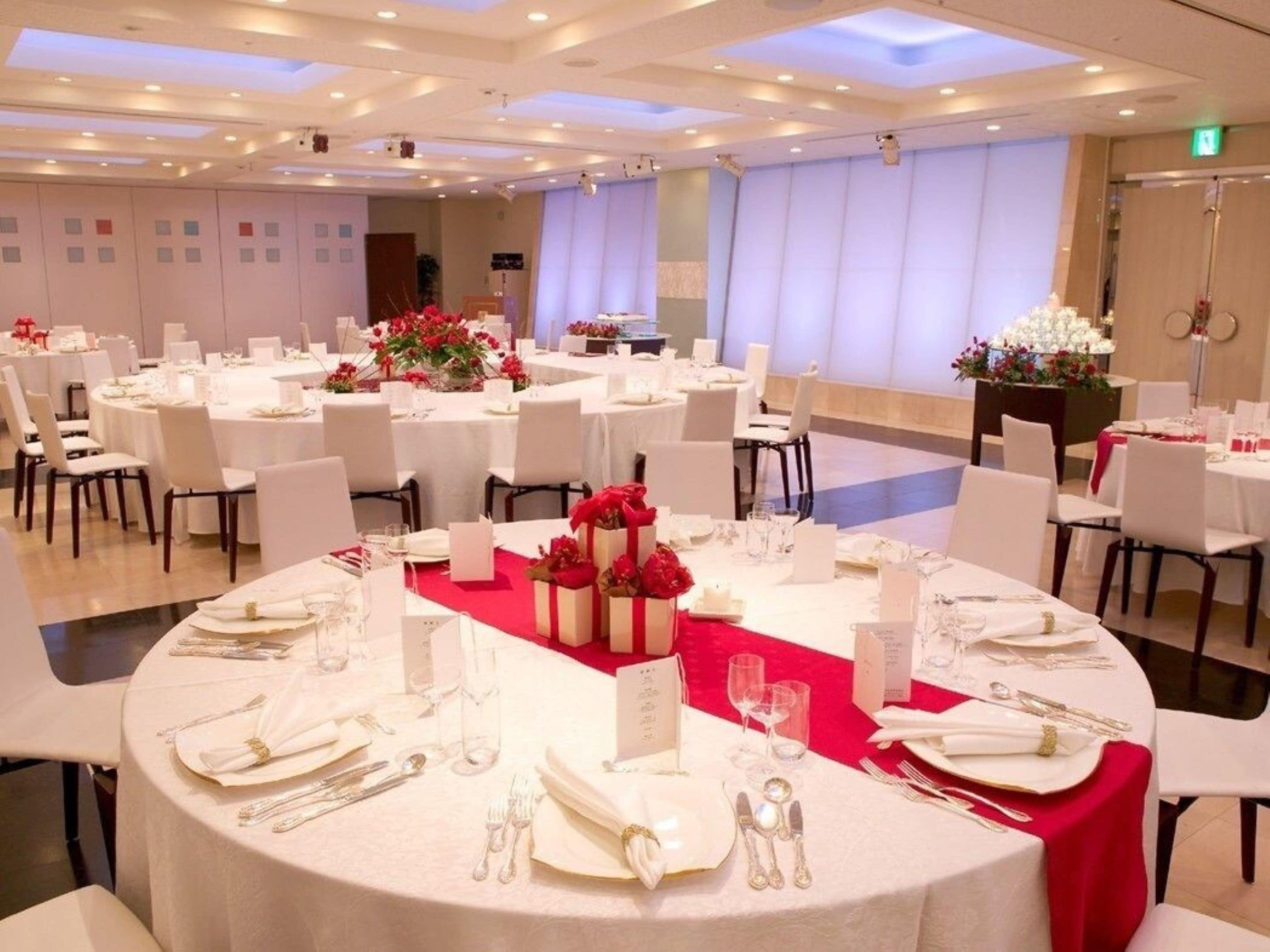A banquet hall with round tables set for dining, white chairs, and red centerpieces.