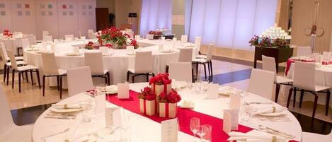 Un salón de banquetes con mesas redondas dispuestas para comer, sillas blancas y centros de mesa rojos.