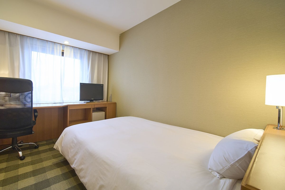 Een hotelkamer met een bed, een bureau met een computer, een stoel, een lamp en een raam met gordijnen.