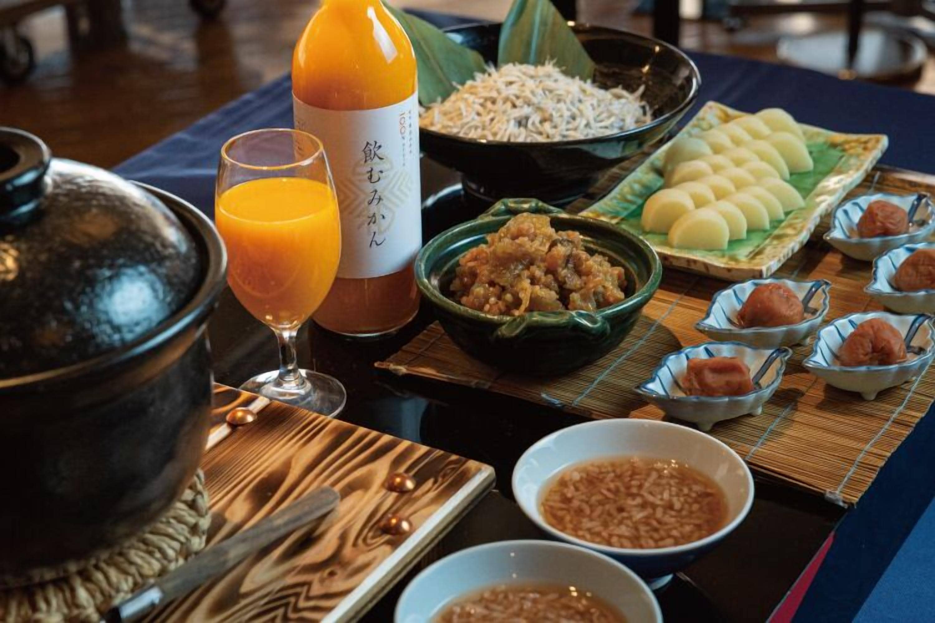 A traditional Japanese meal setup with a pot, bowls of rice, pickled vegetables, and a bottle of orange juice.